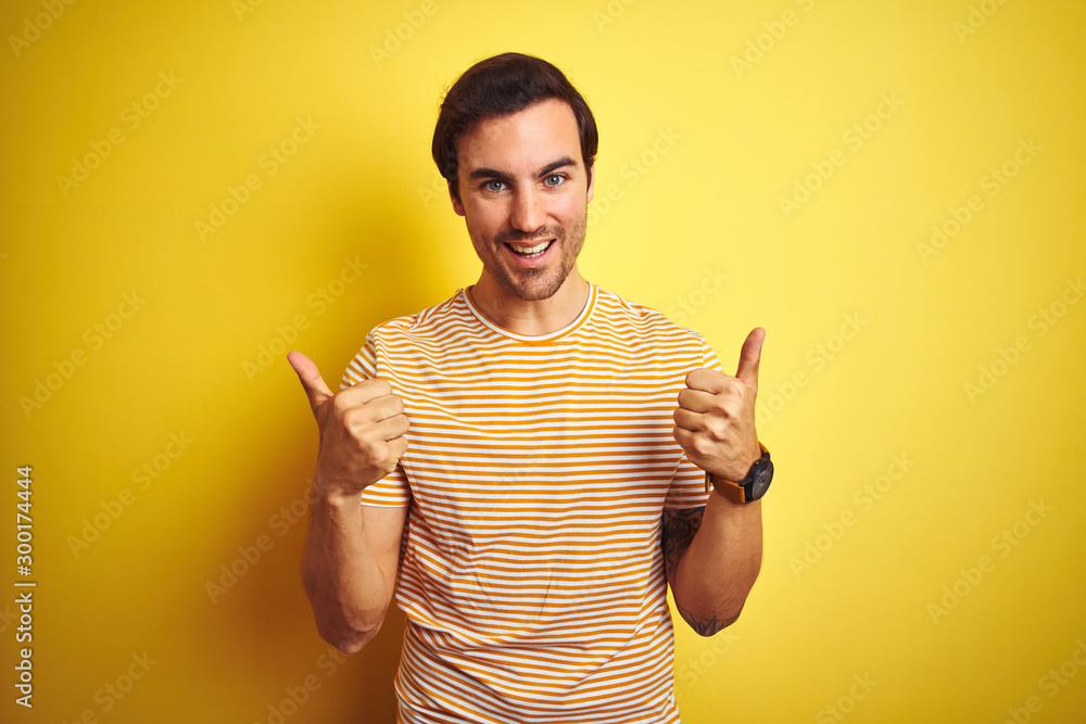 Young handsome man with tattoo wearing striped t-shirt over isolated yellow background success sign doing positive gesture with hand, thumbs up smiling and happy. Cheerful expression Young handsome man with tattoo wearing striped t-shirt over isolated yellow background success sign doing positive gesture with hand, thumbs up smiling and happy. Cheerful expression