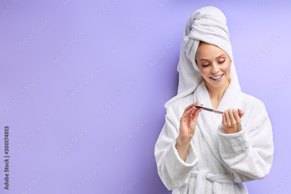 beautiful woman in bathrobe and towel on head cuts nails with nailfile, look at nails. Isolated over purple background beautiful woman in bathrobe and towel on head cuts nails with nailfile, look at nails. Isolated over purple background