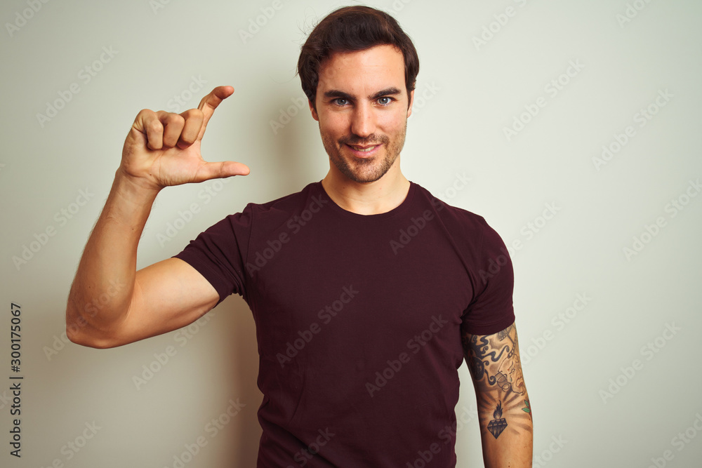 Young handsome man with tattoo wearing purple casual t-shirt over isolated white background smiling and confident gesturing with hand doing small size sign with fingers looking and the camera Young handsome man with tattoo wearing purple casual t-shirt over isolated white background smiling and confident gesturing with hand doing small size sign with fingers looking and the camera