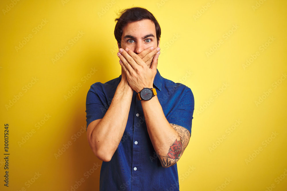 Young handsome man with tattoo wearing casual shirt standing over isolated yellow background shocked covering mouth with hands for mistake. Secret concept. Young handsome man with tattoo wearing casual shirt standing over isolated yellow background shocked covering mouth with hands for mistake. Secret concept.
