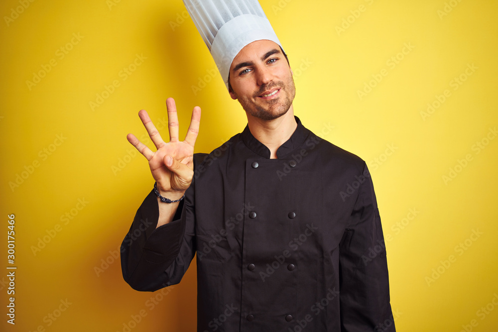 Young chef man wearing uniform and hat standing over isolated yellow background showing and pointing up with fingers number four while smiling confident and happy.