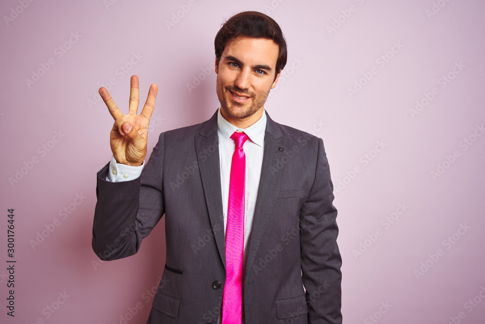 Young handsome businessman wearing suit and tie standing over isolated pink background showing and pointing up with fingers number three while smiling confident and happy. Young handsome businessman wearing suit and tie standing over isolated pink background showing and pointing up with fingers number three while smiling confident and happy.