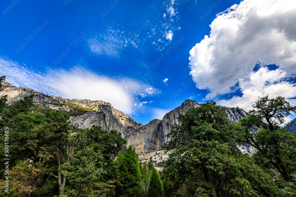 Mostly blue sky with some white clouds, granite mountains and green lush trees in the bottom foreground