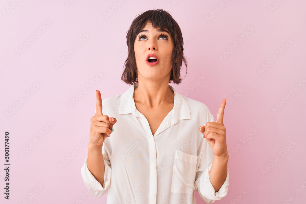 Young beautiful woman wearing white shirt standing over isolated pink background amazed and surprised looking up and pointing with fingers and raised arms. Young beautiful woman wearing white shirt standing over isolated pink background amazed and surprised looking up and pointing with fingers and raised arms.