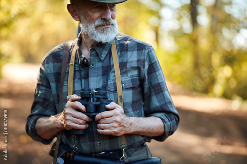 man with grey beard holding binoculars during hunting in autumn forest, looking away. Road background photo