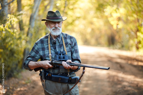 Older hunter man with grey beard holding gun to hunt on birds, ready to shoot. Forest background