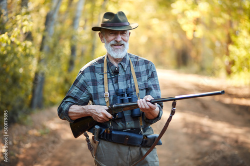 Photographie Portrait of positive hunter man holding gun, stand in forest