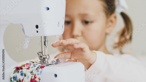 A little blonde girl is attentively sewing a colorful piece of fabric. Hands and a white sewing machine are in focus. Close-up.