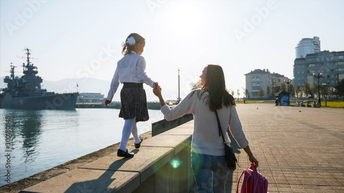 A little girl in school uniform with mother is walking on the railing of the promenade along the sea. A woman is holding her hand, back view.