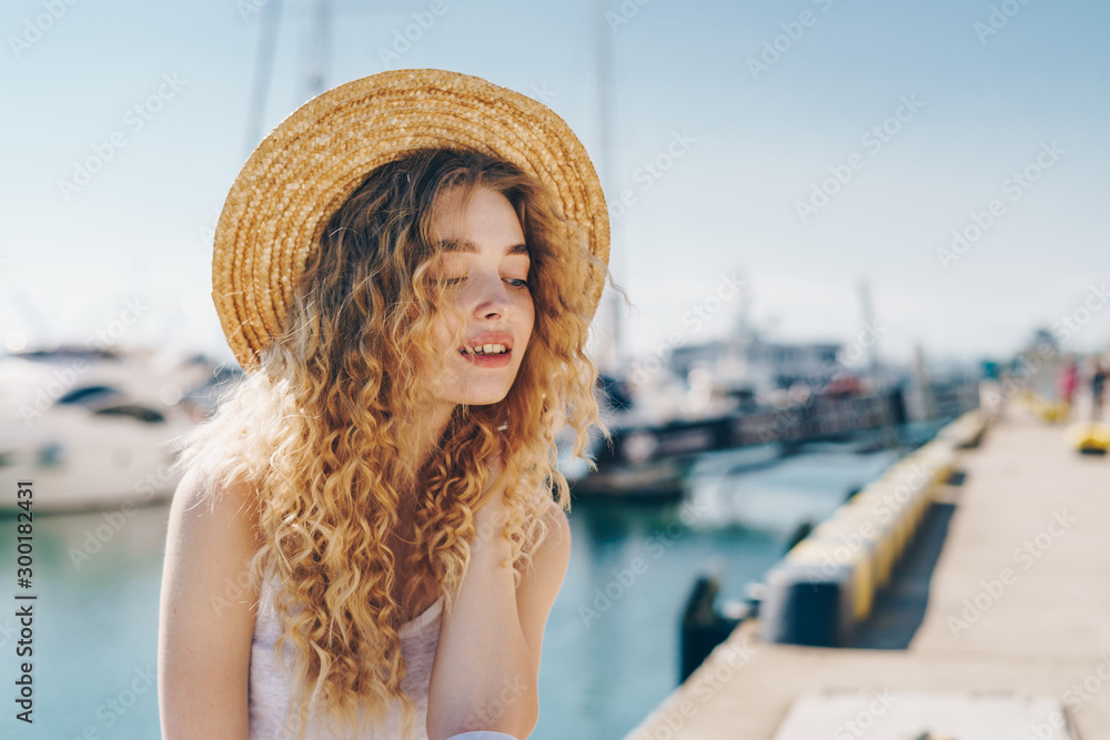 curly tender blonde in a hat on the background of the ships sits on the parapet of the promenade