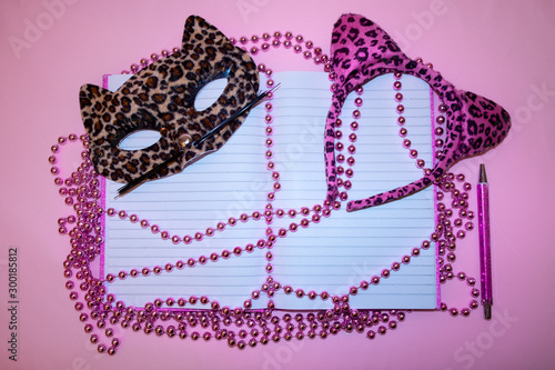 A pink diary and note book on pink background. Pink pearls on long strings, a cute masquerade cat mask and cat ears in leopard pattern.