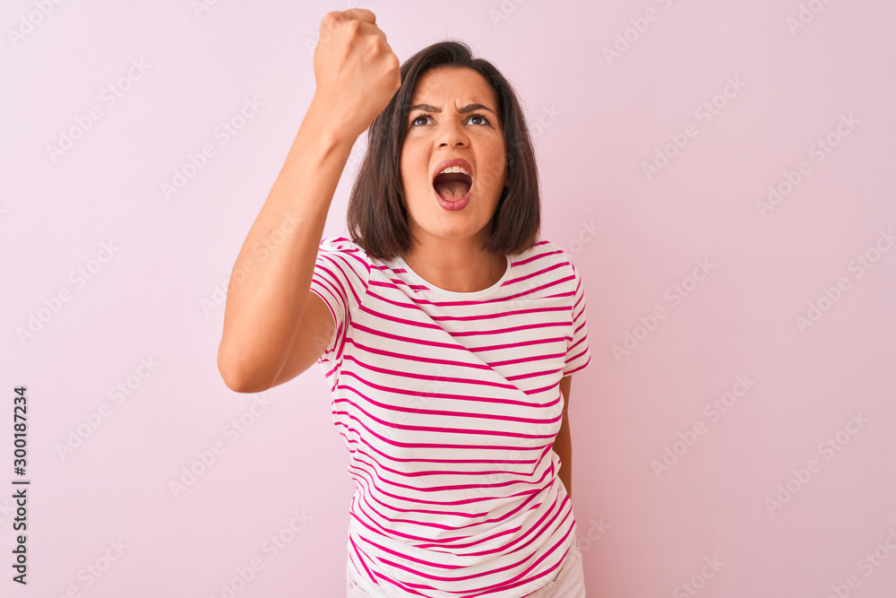 Young beautiful woman wearing striped t-shirt standing over isolated pink background angry and mad raising fist frustrated and furious while shouting with anger. Rage and aggressive concept. Young beautiful woman wearing striped t-shirt standing over isolated pink background angry and mad raising fist frustrated and furious while shouting with anger. Rage and aggressive concept.
