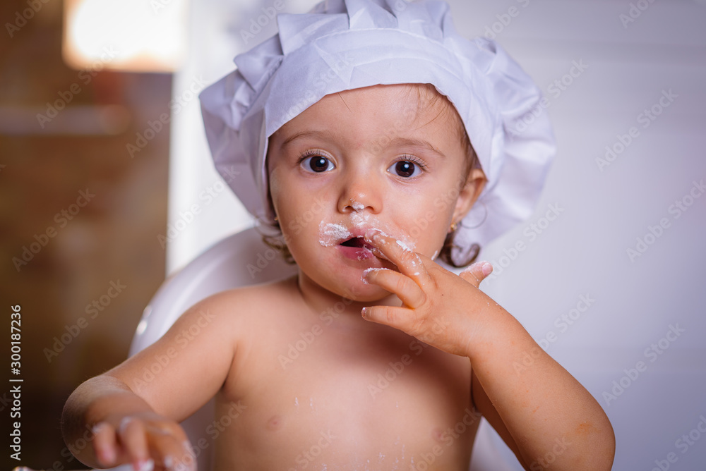 A sweet small chef cook in a white culinary cap the eating flour from fingers. Happy girl bullies in the kitchen. A sweet small chef cook in a white culinary cap the eating flour from fingers. Happy girl bullies in the kitchen.