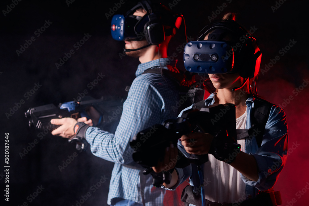 Man and woman with vr guns and headsets standing isolated black background with red neon backlit. entertainmnet, copy space