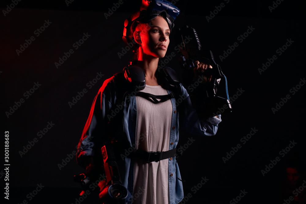 Attractive caucasian girl wearing special equipment, holding weapon for virtual game. Stand posing isolated over black background