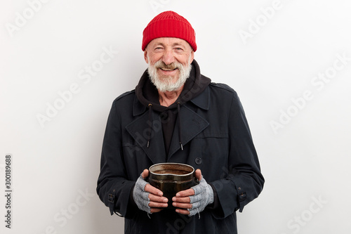 Mature smiling bum in black coat and red cap on head holding iron can for raising money. Isolated over white background