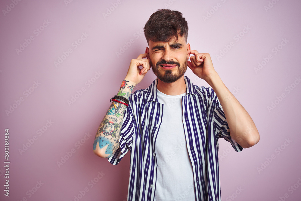Obraz premium Young man with tattoo wearing striped shirt standing over isolated pink background covering ears with fingers with annoyed expression for the noise of loud music. Deaf concept.