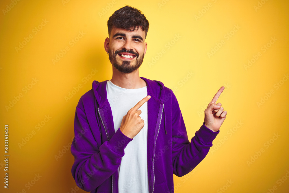 Young man with tattoo wearing sport purple sweatshirt over isolated yellow background smiling and looking at the camera pointing with two hands and fingers to the side. Young man with tattoo wearing sport purple sweatshirt over isolated yellow background smiling and looking at the camera pointing with two hands and fingers to the side.