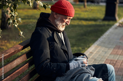 Grey bearded man without shelter sitting in street clothes on bench, without food and money. Drooping man lowered his head down. Side view on homeless person in coat and red cap