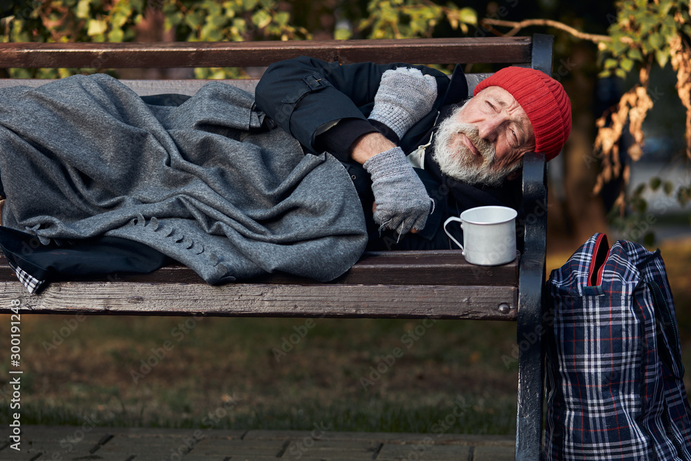 Penniless senior man lying on park bench, trembling from the cold. Cup for collecting money, coins next to him. Penniless senior man lying on park bench, trembling from the cold. Cup for collecting money, coins next to him.