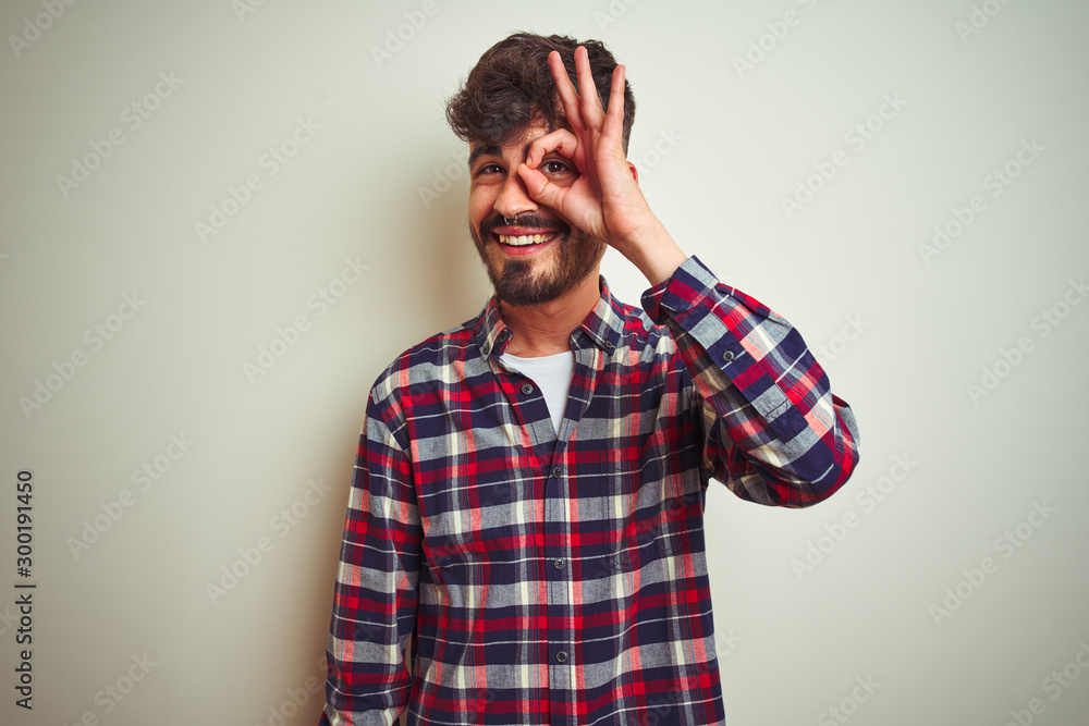 Young man wearing casual shirt standing over isolated white background doing ok gesture with hand smiling, eye looking through fingers with happy face. Young man wearing casual shirt standing over isolated white background doing ok gesture with hand smiling, eye looking through fingers with happy face.