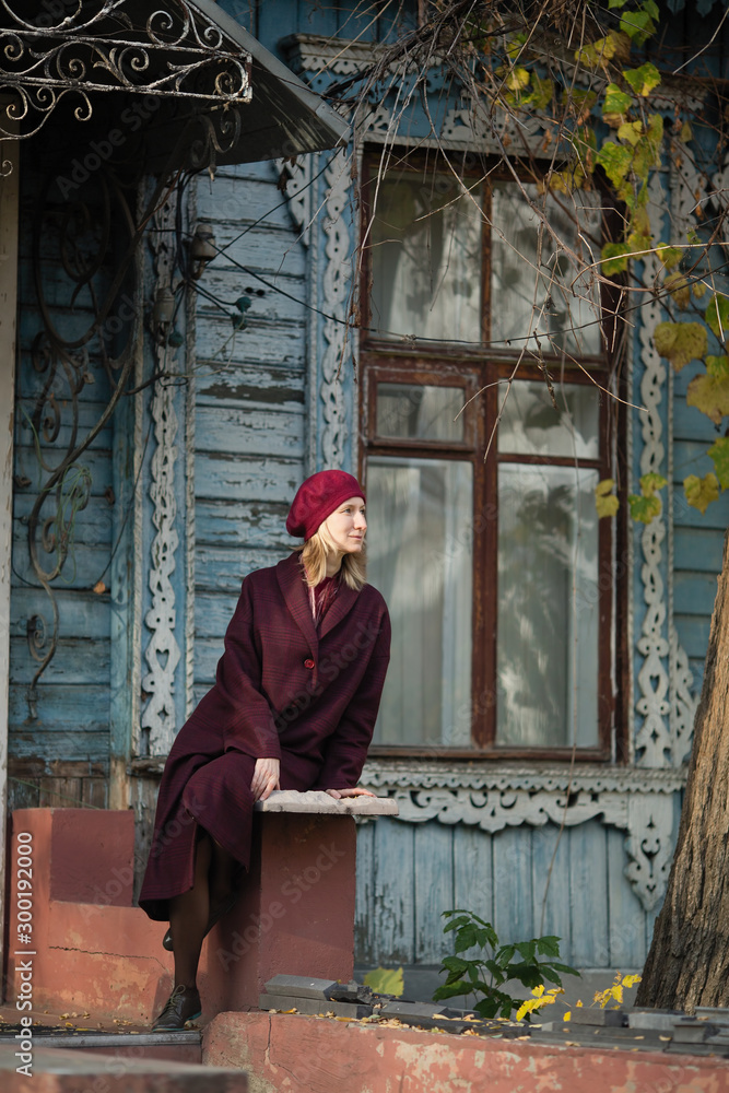 Blonde woman in a burgundy coat and beret sitting on porch of old wooden house.