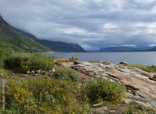 view on Akkajaure lake with green mountains, rock boulders, bushes, birch tree and dramatic clouds photo