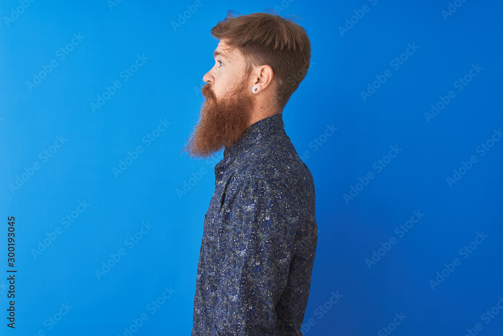 Young redhead irish man wearing floral summer shirt standing over isolated blue background looking to side, relax profile pose with natural face with confident smile.