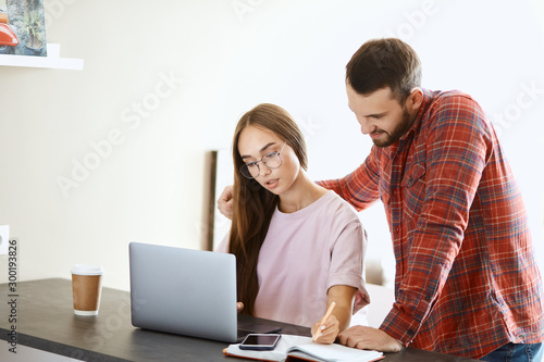 Concentrated pretty woman sits at table, works at laptop computer, stares at screen, writes down on paper, holds pencil, spending time at home together with husband