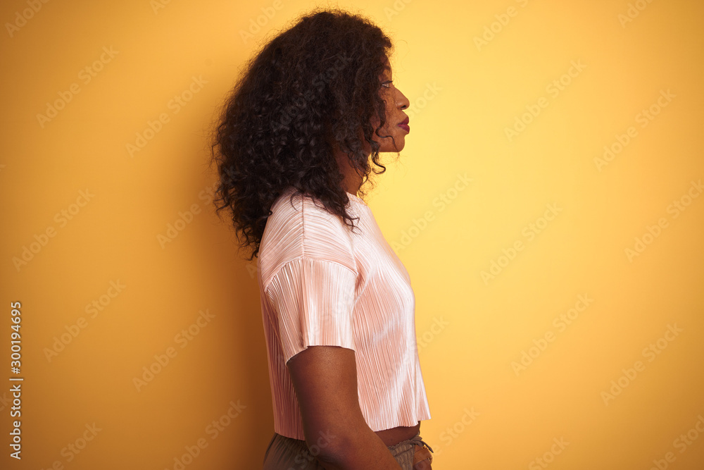 African american woman wearing pink t-shirt standing over isolated yellow background looking to side, relax profile pose with natural face with confident smile. African american woman wearing pink t-shirt standing over isolated yellow background looking to side, relax profile pose with natural face with confident smile.