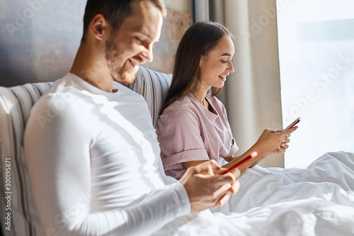 Positive atmosphere, saturday morning, sun shining brightly, young people ignore live communication and looking at cell phone screens, expressing happiness, chatting in web blog, family concept