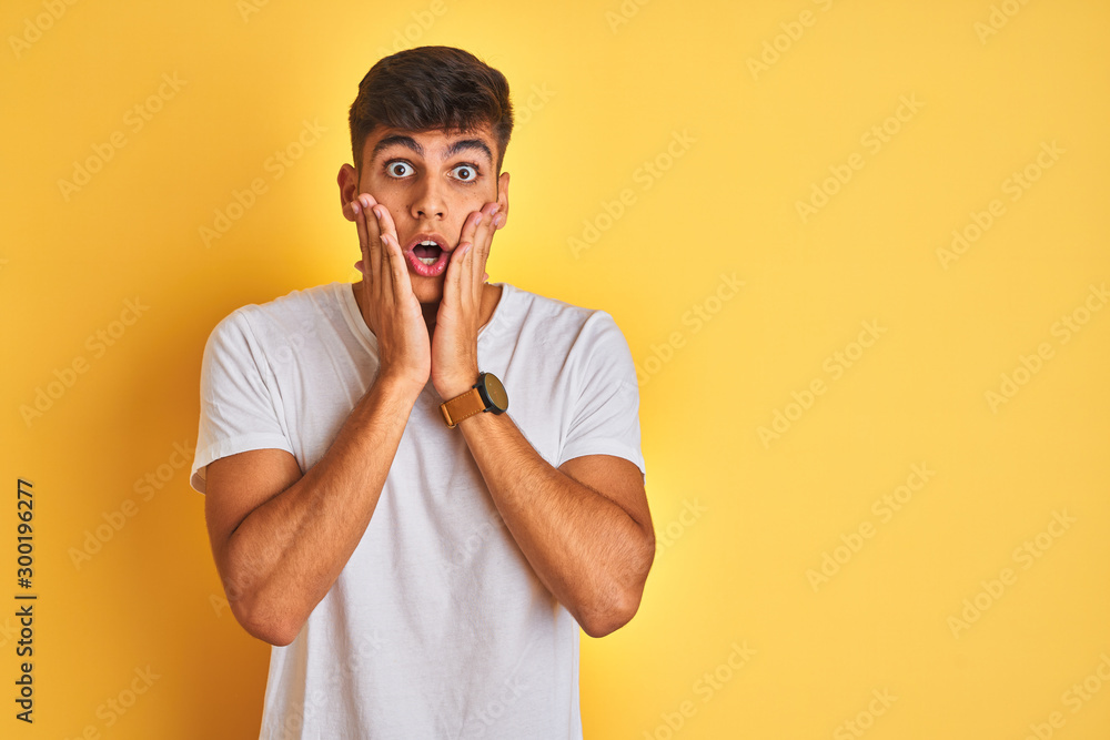 Young indian man wearing white t-shirt standing over isolated yellow background afraid and shocked, surprise and amazed expression with hands on face Young indian man wearing white t-shirt standing over isolated yellow background afraid and shocked, surprise and amazed expression with hands on face
