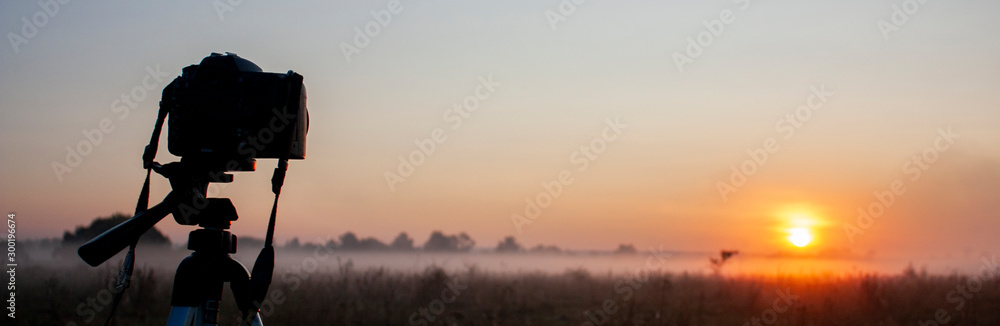 Silhouette of a camera on a tripod early in the morning. Fog on the field and orange sun over the horizon. Summer morning on the field.