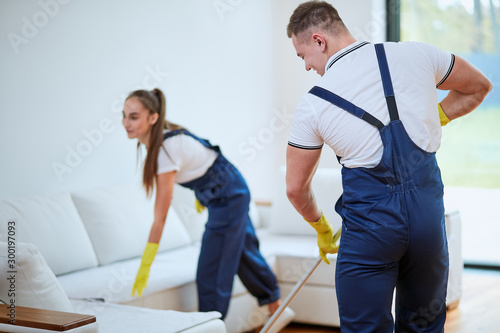 Two janitors washing floor near sofa. man and woman in uniform, overalls and yellow rubber gloves with mops.