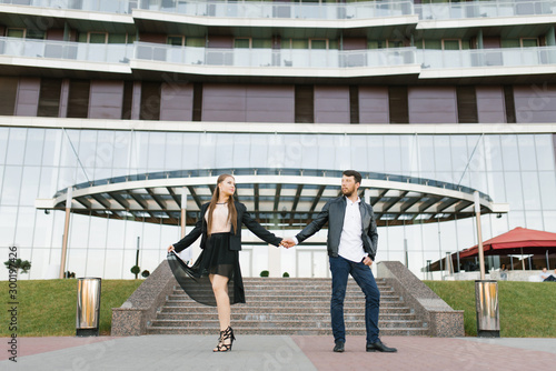 A guy and a girl in dark clothes hold hands on the background of a glass building