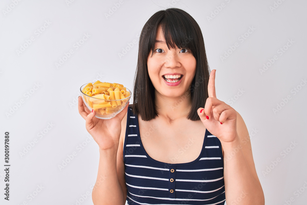 Young beautiful Chinese woman holding bowl with macaroni over isolated white background surprised with an idea or question pointing finger with happy face, number one Young beautiful Chinese woman holding bowl with macaroni over isolated white background surprised with an idea or question pointing finger with happy face, number one