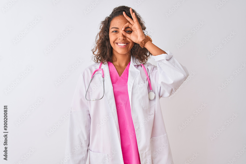 Young brazilian doctor woman wearing stethoscope standing over isolated white background doing ok gesture with hand smiling, eye looking through fingers with happy face. Young brazilian doctor woman wearing stethoscope standing over isolated white background doing ok gesture with hand smiling, eye looking through fingers with happy face.