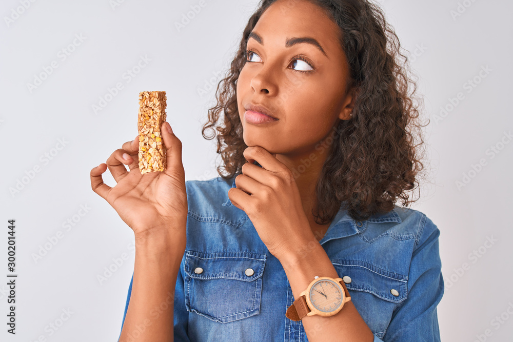Young brazilian woman eating granola bar standing over isolated white background serious face thinking about question, very confused idea Young brazilian woman eating granola bar standing over isolated white background serious face thinking about question, very confused idea