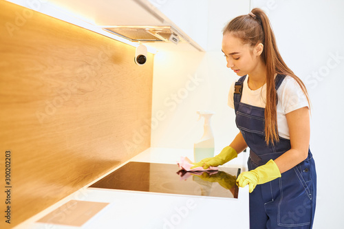 Side view on young cleaner wearing yellow rubber gloves wipes sensory stove, white interior kitchen