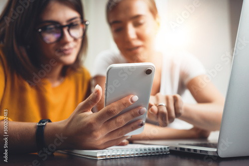Close-up of female hands hold modern smartphone at her workplace at office. Group of posotive woman working together and using digital gadgets at open space