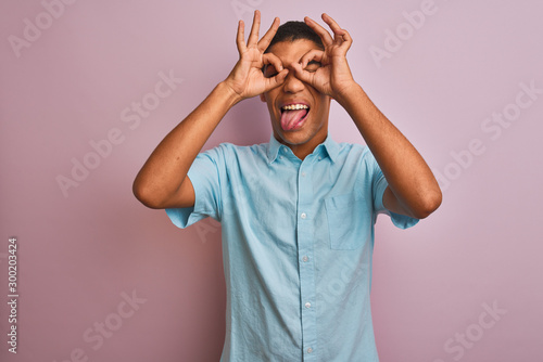 Young handsome arab man wearing blue shirt standing over isolated pink background doing ok gesture like binoculars sticking tongue out, eyes looking through fingers. Crazy expression.