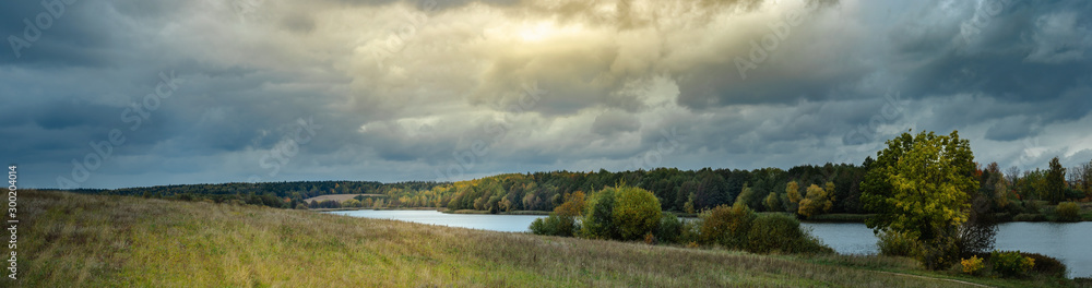 a wide panoramic view of the river with coastal meadows on a hillside, forests and a dramatic stormy dark sky with a sunny glow