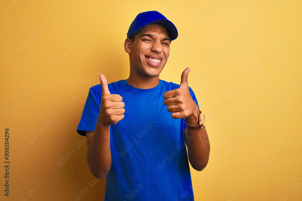 Young handsome arab delivery man standing over isolated yellow background success sign doing positive gesture with hand, thumbs up smiling and happy. Cheerful expression and winner gesture.