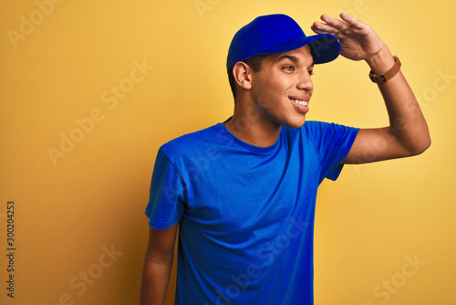 Young handsome arab delivery man standing over isolated yellow background very happy and smiling looking far away with hand over head. Searching concept.