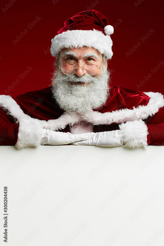 Naklejka premium Portrait of classic Santa Claus smiling gleefully at camera while posing over blank white sign, copy space