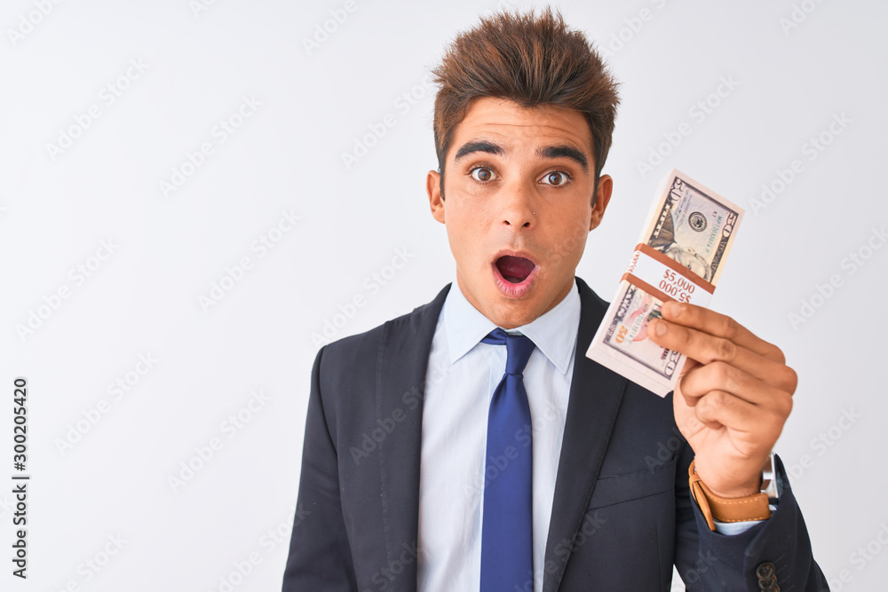 Young handsome businessman wearing suit holding dollars over isolated white background scared in shock with a surprise face, afraid and excited with fear expression Young handsome businessman wearing suit holding dollars over isolated white background scared in shock with a surprise face, afraid and excited with fear expression
