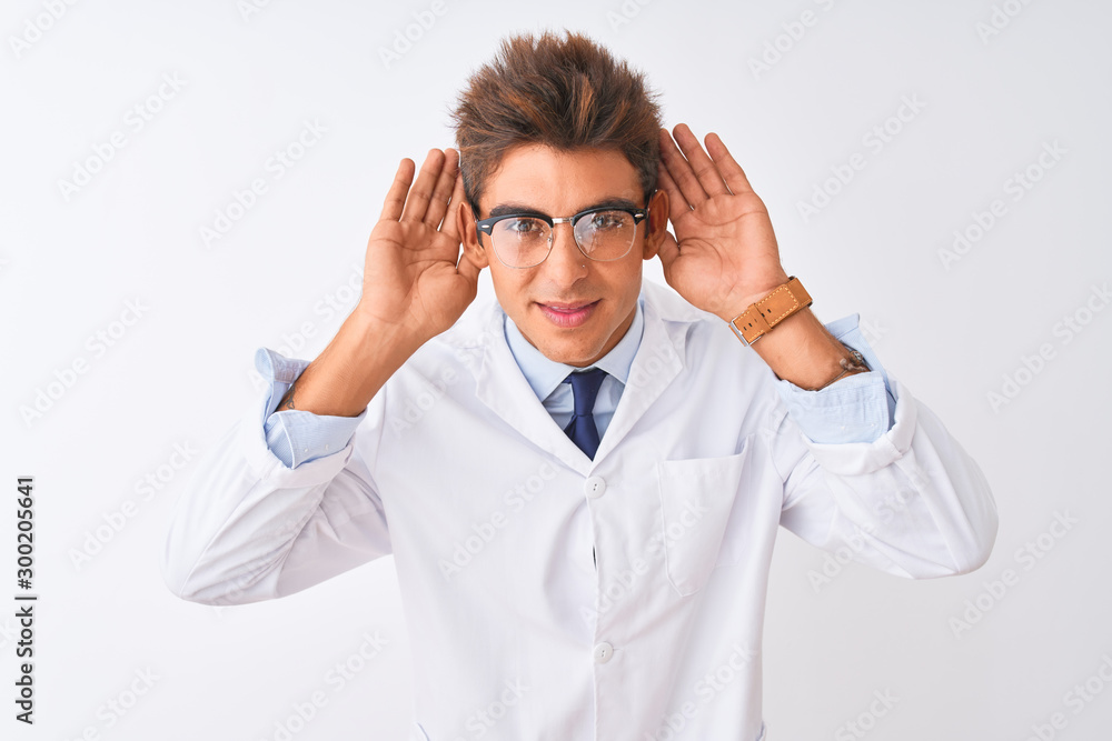 Young handsome sciencist man wearing glasses and coat over isolated white background Trying to hear both hands on ear gesture, curious for gossip. Hearing problem, deaf Young handsome sciencist man wearing glasses and coat over isolated white background Trying to hear both hands on ear gesture, curious for gossip. Hearing problem, deaf