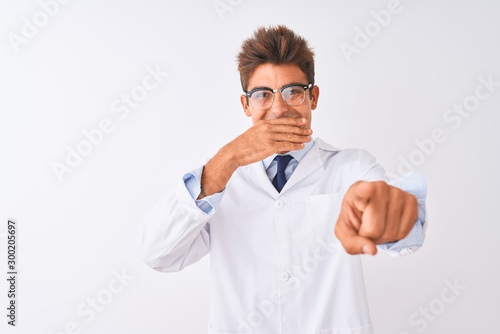 Young handsome sciencist man wearing glasses and coat over isolated white background laughing at you, pointing finger to the camera with hand over mouth, shame expression © Krakenimages.com