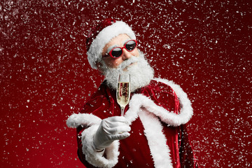 Waist up portrait of cool Santa holding champagne glass while standing over red background with snow falling, copy space
