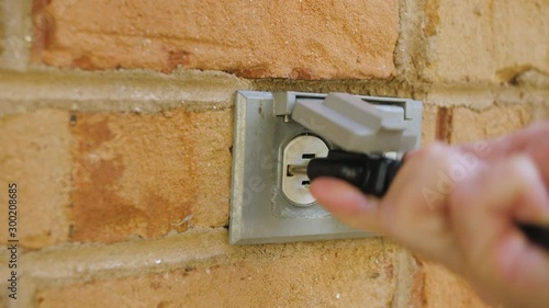 Man plugging in a power cord and then unplugging it from an outdoor electrical outlet. Dual covered outdoor power sockets on brick wall. Black extension cord plug inserted into a wall socket.